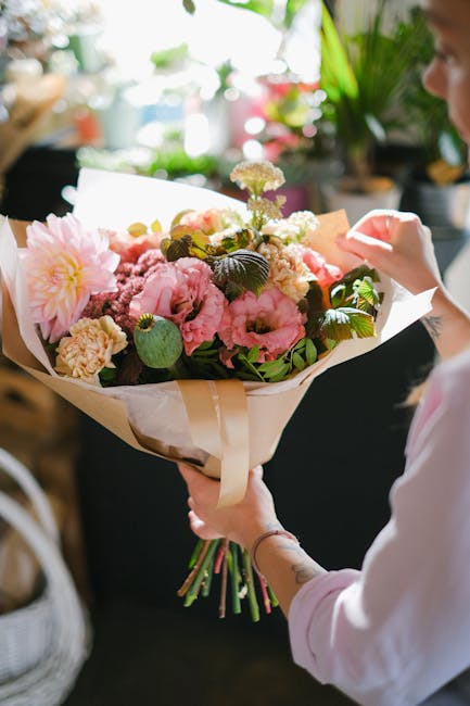 A person holding a large floral bouquet featuring soft pink peonies, cream-colored carnations, and delicate white astilbes, accented with lush green foliage. The bouquet is wrapped in elegant cream an