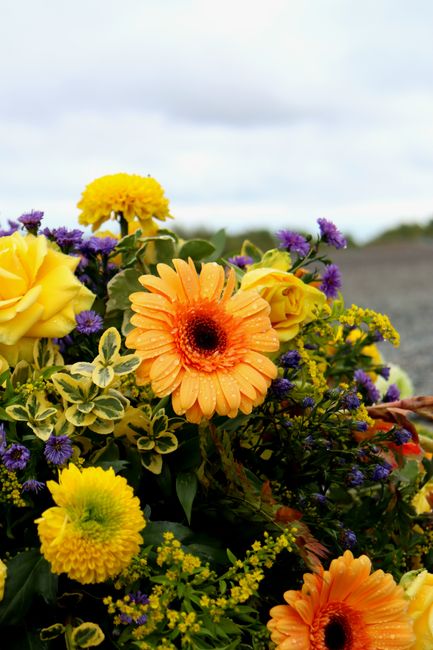 A vibrant floral arrangement featuring large yellow roses, bright orange gerbera daisies, and small purple flowers, all with fresh, dewy petals and sturdy green stems. The bouquet is densely packed wi