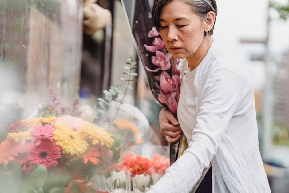 A woman with short gray hair and a focused expression is selecting fresh flowers at an outdoor flower market. She is holding a bouquet of vibrant pink orchids close to her chest, while her other hand 