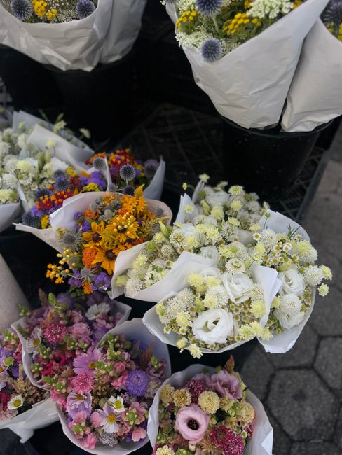A display of fresh floral bouquets at a flower market, featuring a variety of seasonal blooms arranged in small, white wrapping paper. The arrangements include vibrant orange sunflowers, purple daisie